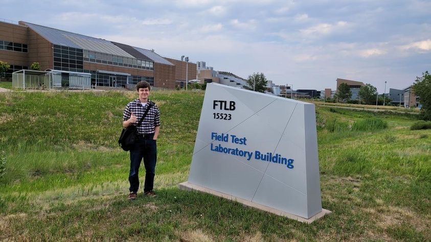 Nathan stands beside an office sign outside on the grass. 