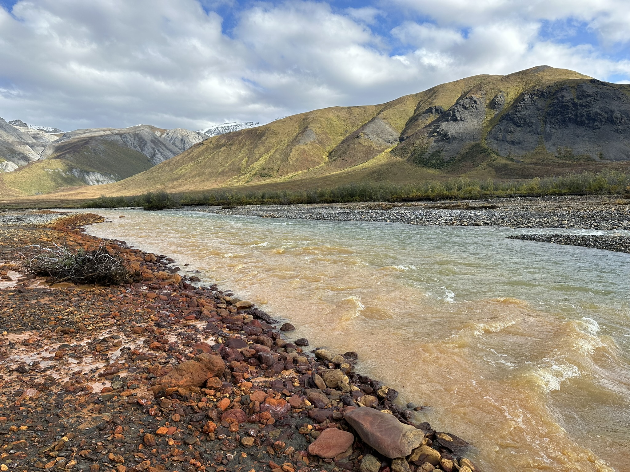 River with milky turquoise water flowing past red rocky bank toward grassy, rounded mountains