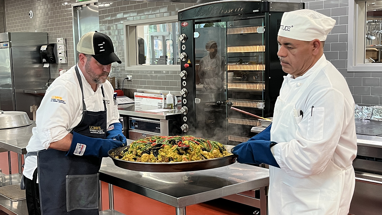 A chef and food service worker transfer a large pan of paella to a serving counter