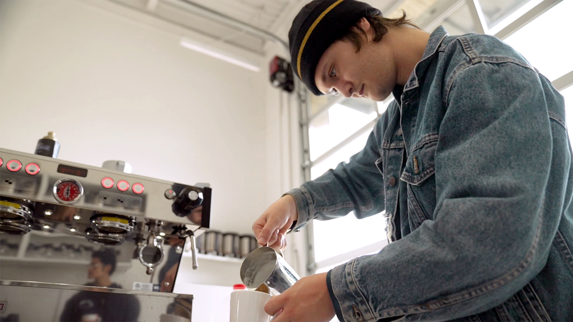 Student Keegan Thompson, wearing a dark beanie and blue denim jacket, pours foam into a white mug.