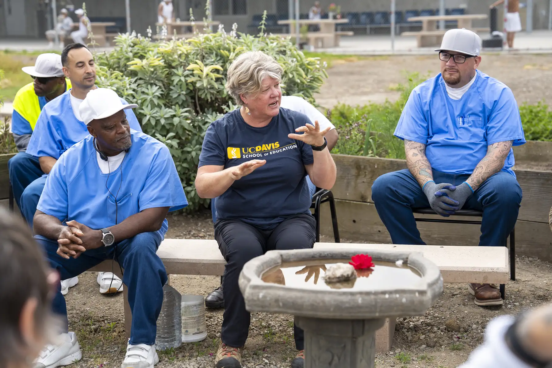 Group seated outdoors around a low planter; a woman gestures while others listen.