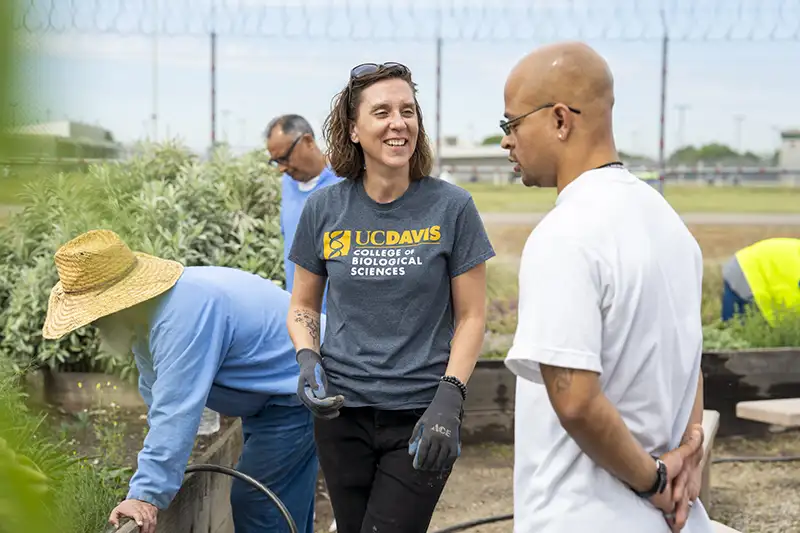 Smiling woman in UC Davis shirt talks with man as others garden outdoors.
