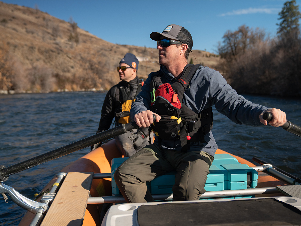 A closeup of two men in a kayak on a river, both wearing sunglasses and caps.