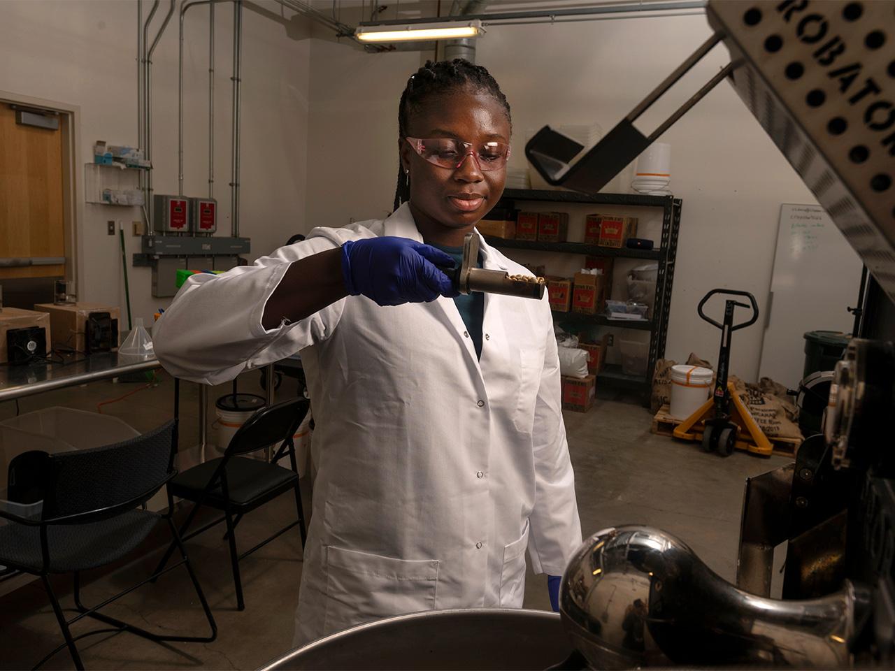 A woman wearing lab gear examines roasted coffee beans in a small tray.