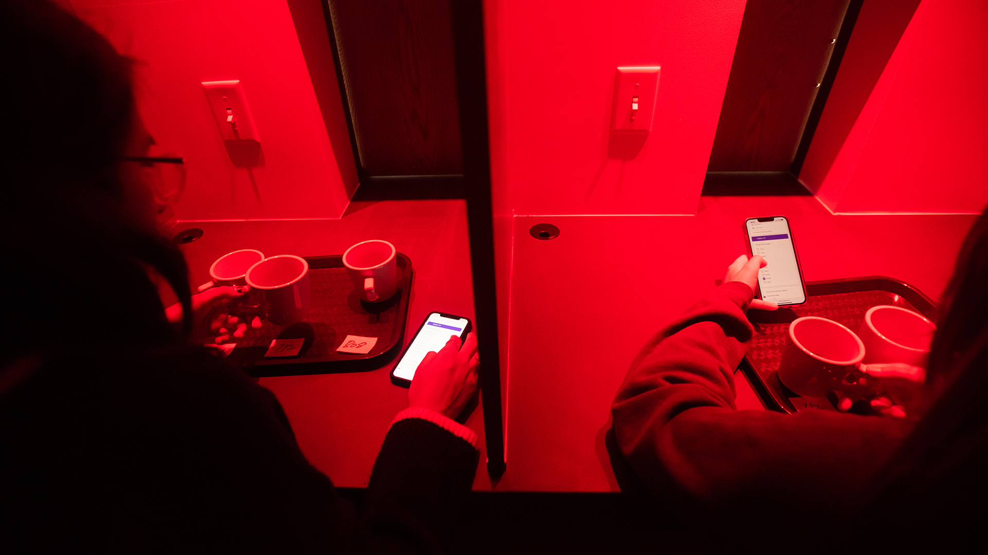 Two students sit in adjacent sensory booths, scrolling on their phones to check boxes beside different flavor descriptors. In front of each student is a tray holding three coffee mugs. In front of each mug is a three-digit, hand-written number on a piece of paper. The photo is lit only by red light.