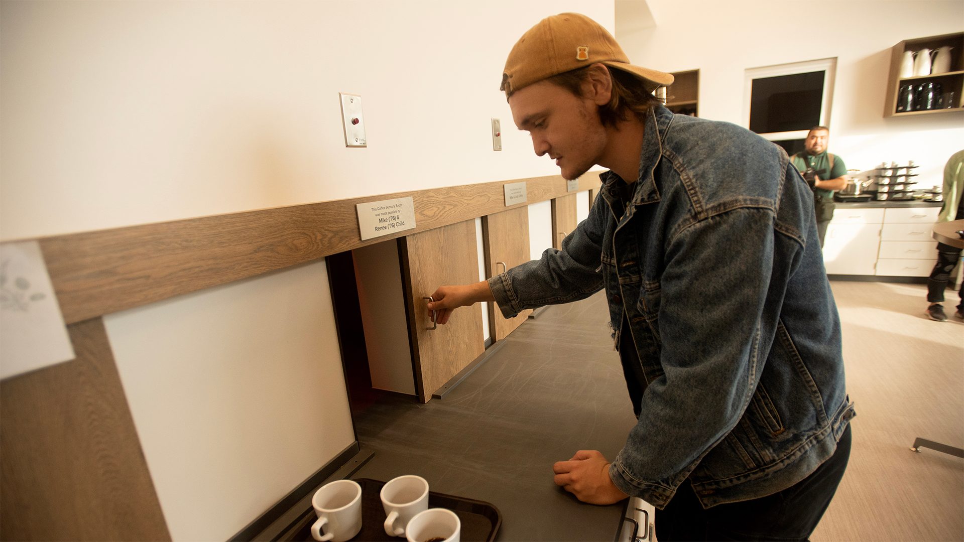 Keegan Thompson, in a backward mustard baseball cap and blue denim jacket, slides open a wooden service door in a white wall, about to deliver the tray with three filled coffee mugs on the dark countertop beside him.