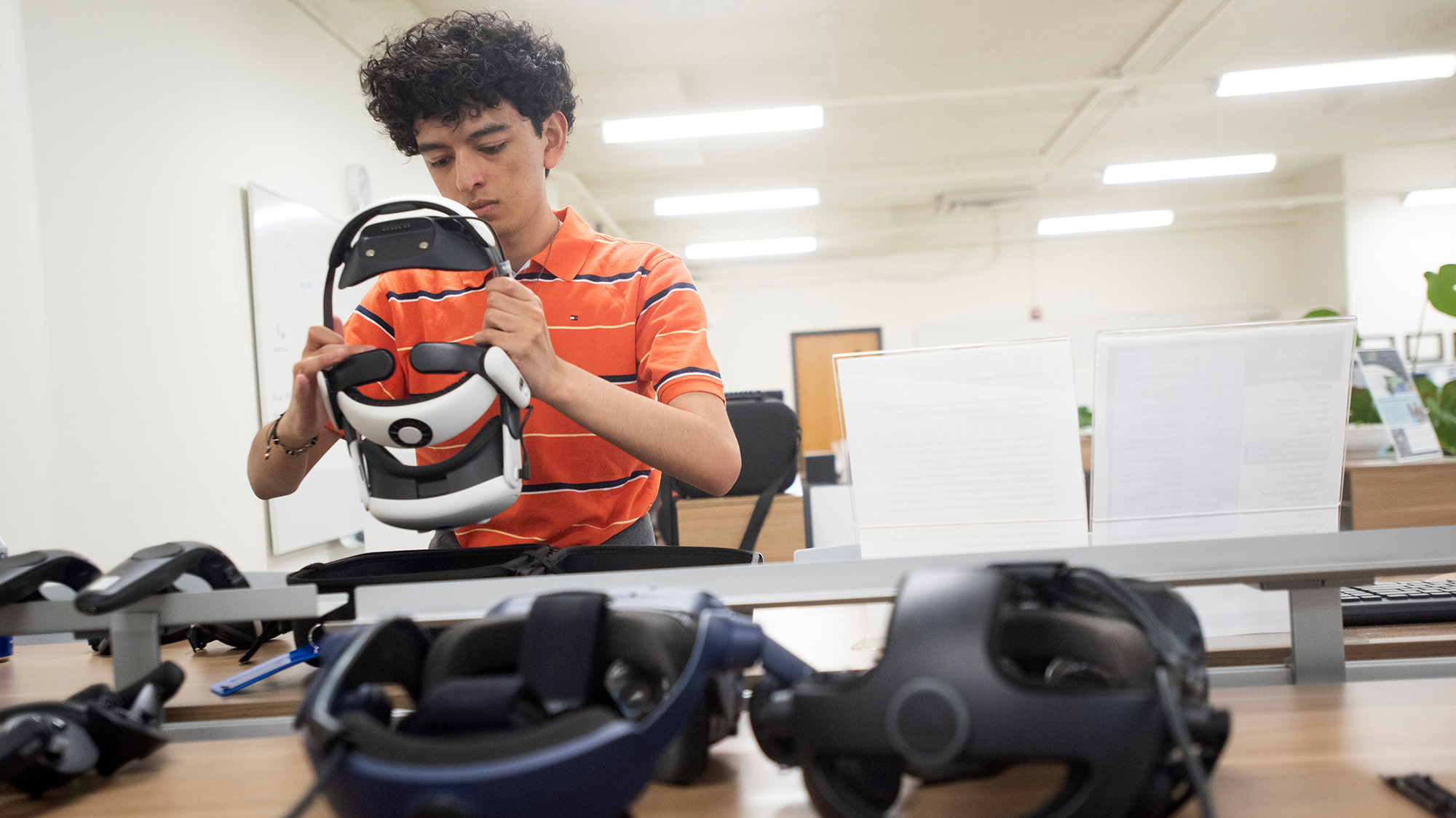 Young person in orange shirt adjusting a VR headset in a tech lab, multiple headsets on table