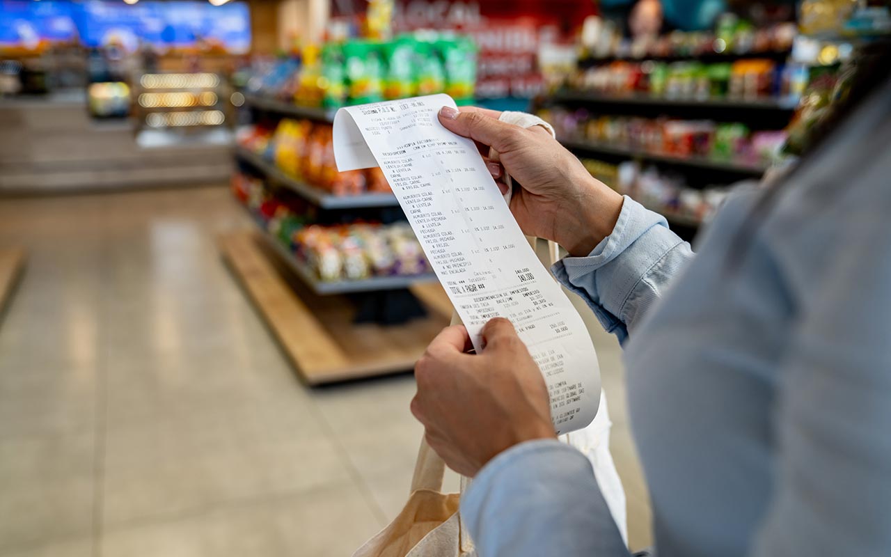 Closeup photo of a woman's hands holding a store receipt to read it. Grocery store shelves are in the background.