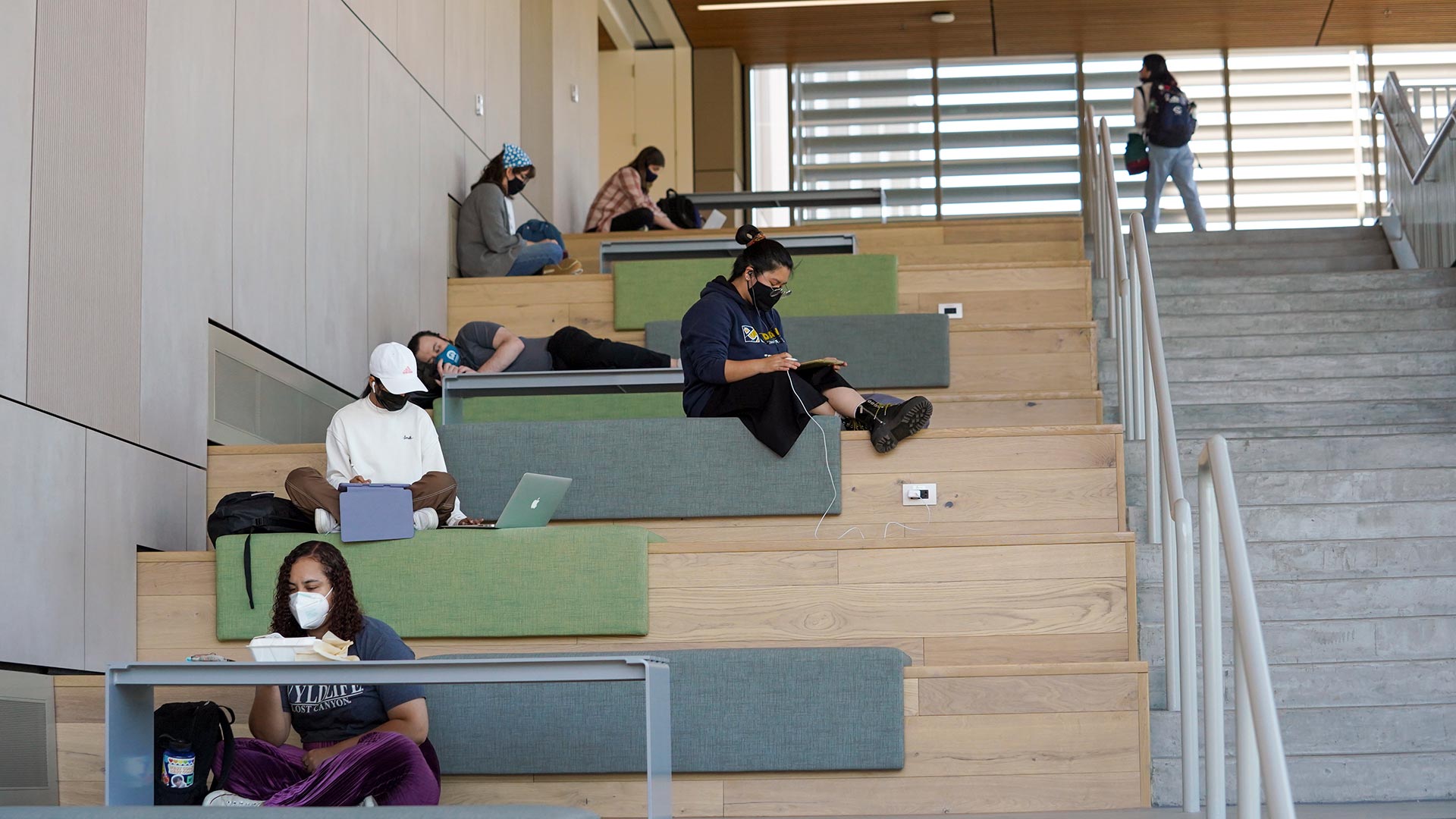 Photo of a concrete staircase on the right with large wooden steps on the left. Students sit in various places along the wooden steps studying.