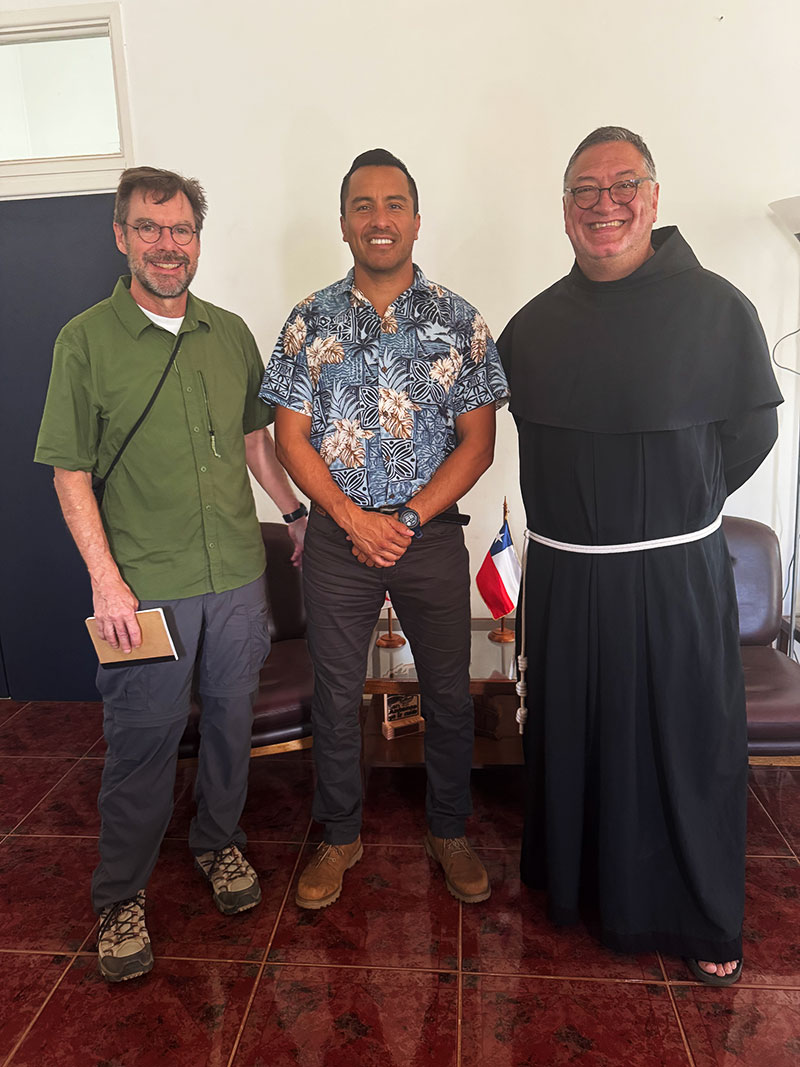 Three men stand for a portrait. One is wearing a green shirt, the next is wearing a flower pattern shirt and the third is wearing Franciscan robes.