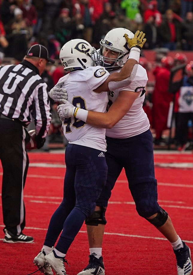 Two Aggie football players hug on red field.