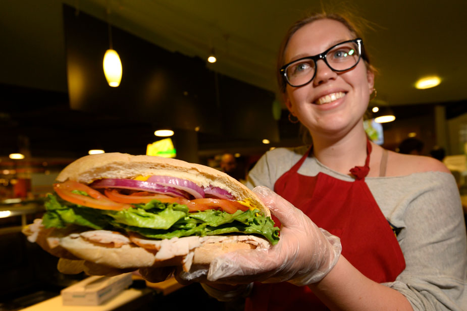 Young woman holds sandwich toward camera.