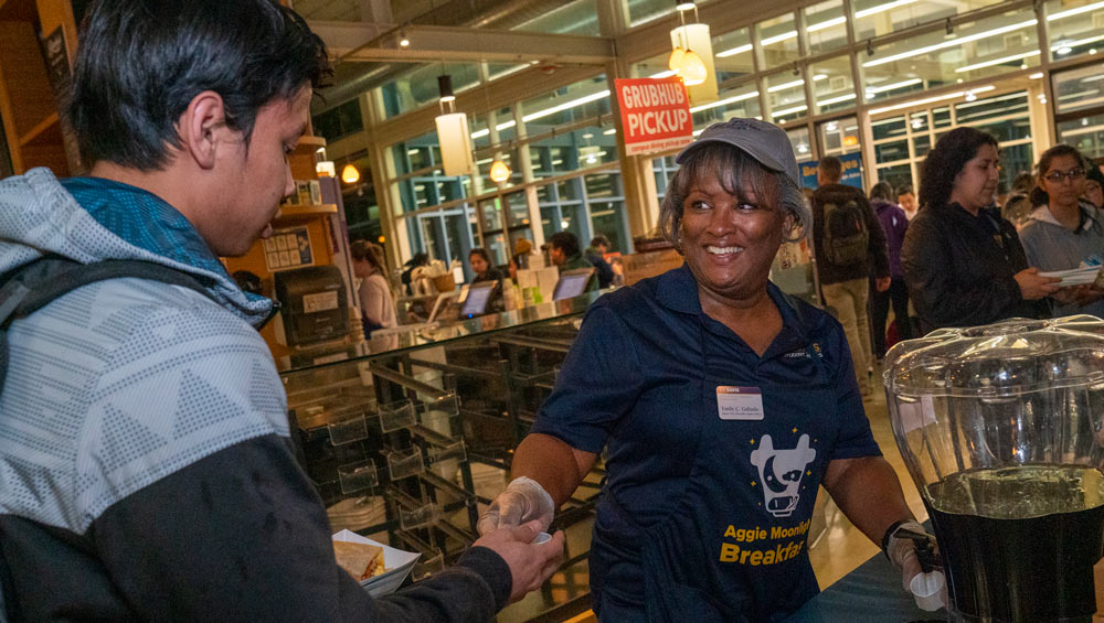 Emily Galindo in Aggie blue apron, smiling, serving a student at Moonlight Breakfast.