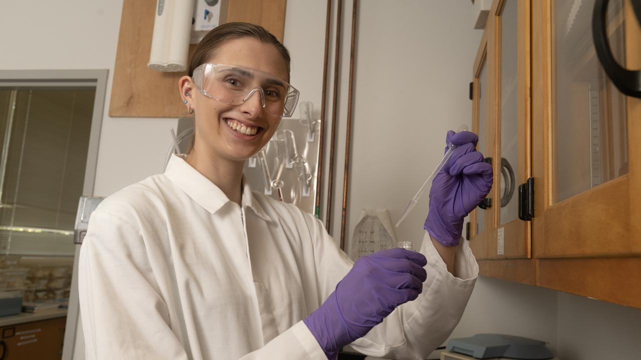 In a white lab coat and goggles, Brooke Morey works in a research lab