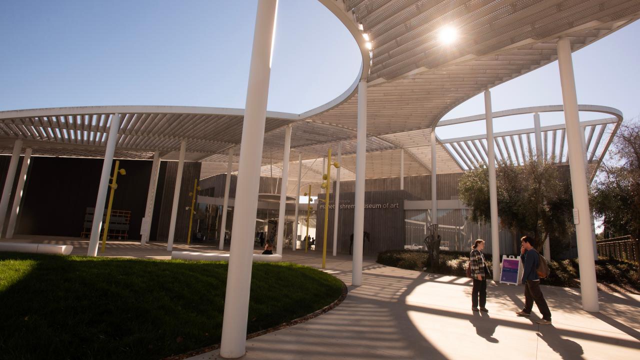 Sunlit modern open-air courtyard with curved wooden canopy, white columns, and two people walking