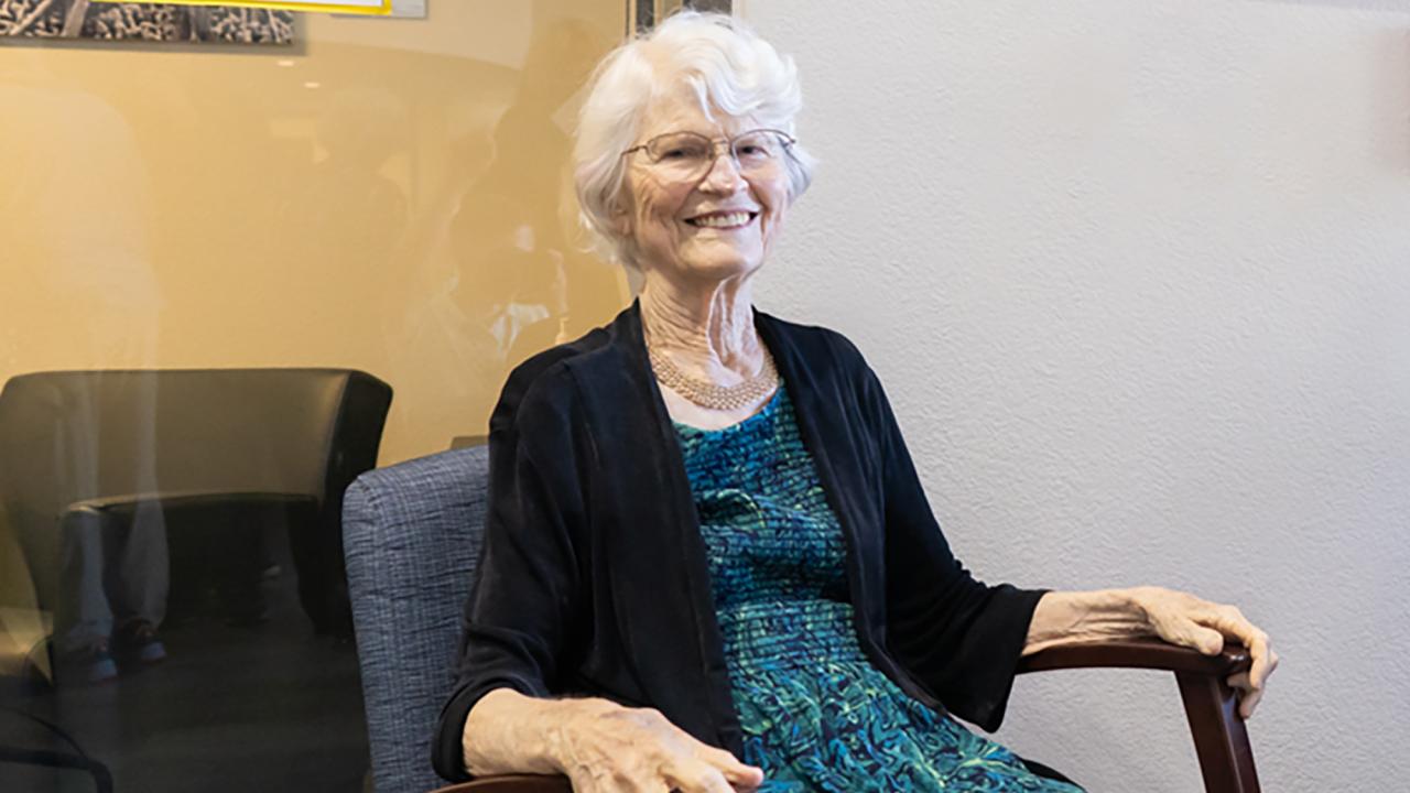 Smiling woman with white hair seated in wooden chair, teal dress