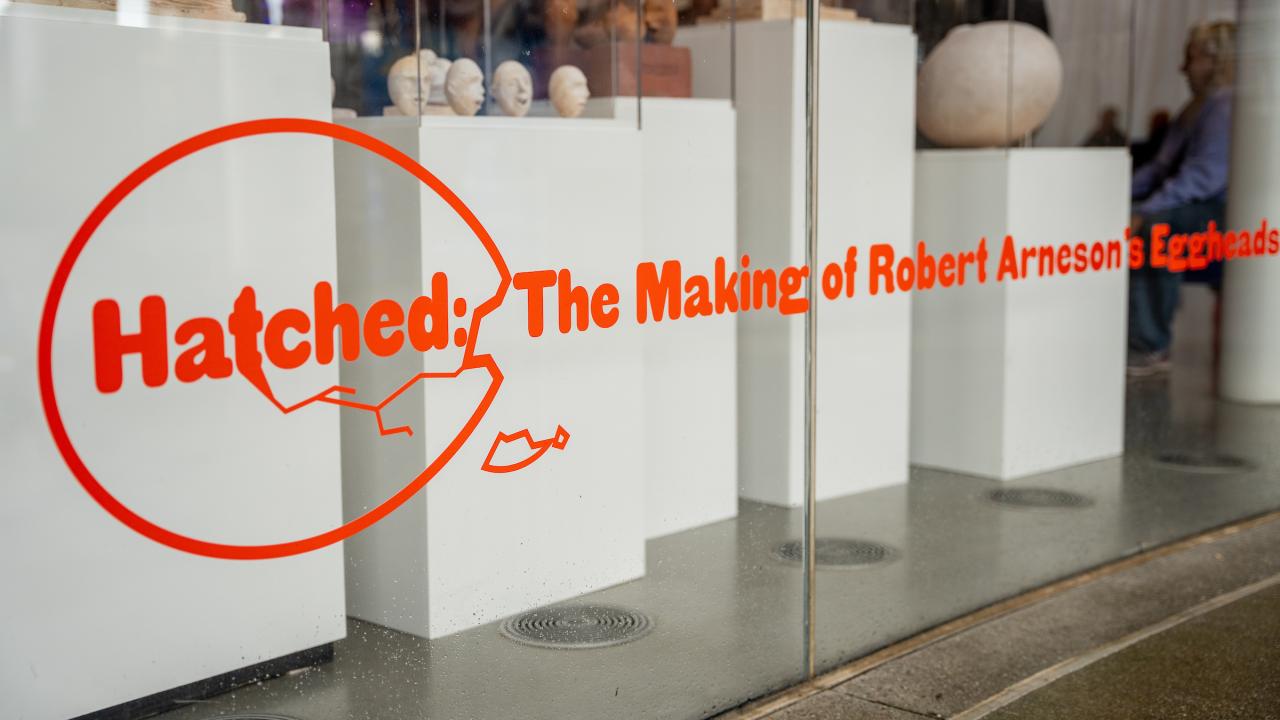 Red letters on a window display announce "Hatched" an art installation inside the lobby of the Manetti Shrem Museum. The photo showcases the exterior window and the small maquette sculptures that modeled Robert Arneson's Egghead sculptures.