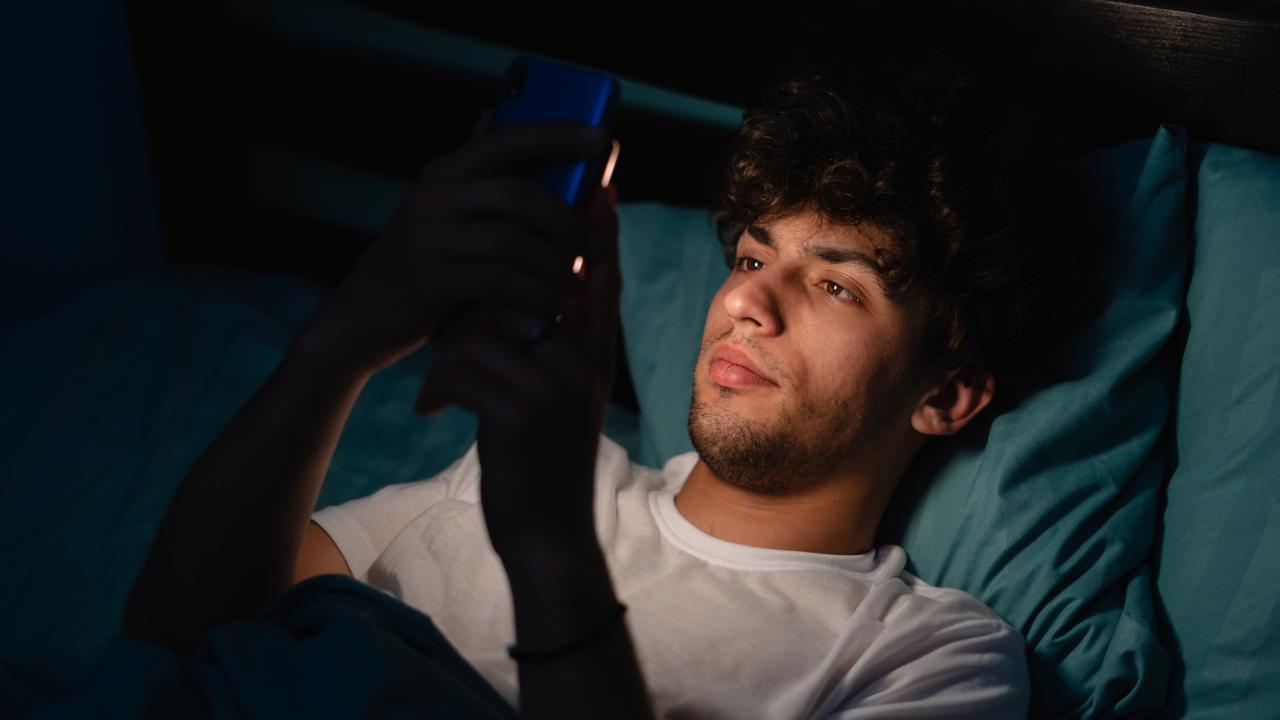Young man in bed, looking at his phone in a dimly lit room.