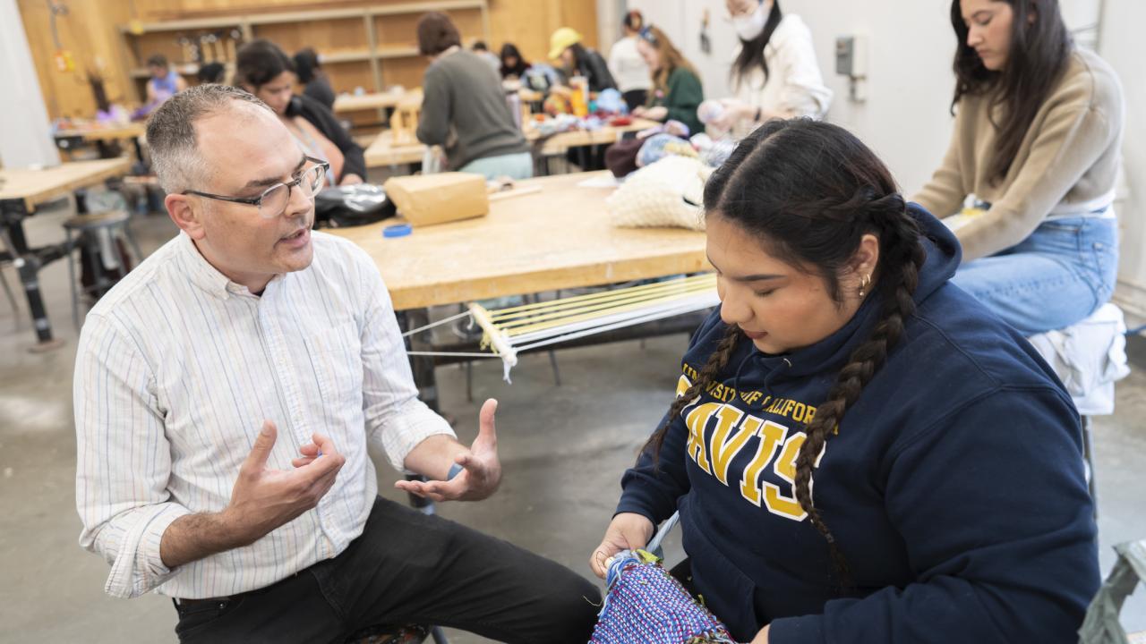 Male instructor speaking to student knitting purple yarn in a busy classroom workshop