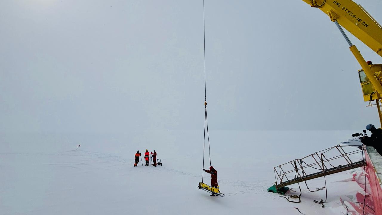 three researchers in coats and a yellow robotic glider are on the ice and snow in Antarctica next to a yellow crane
