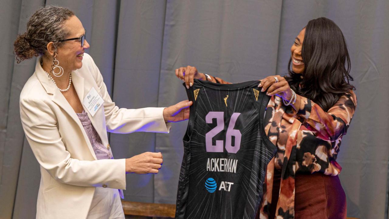 Two women smiling hold a black No.26 basketball jersey on stage.
