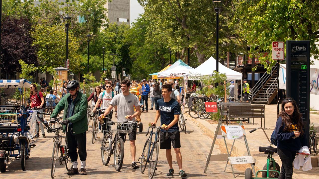 People walk their bicycles down Third Street, with views of UC Davis in the background.