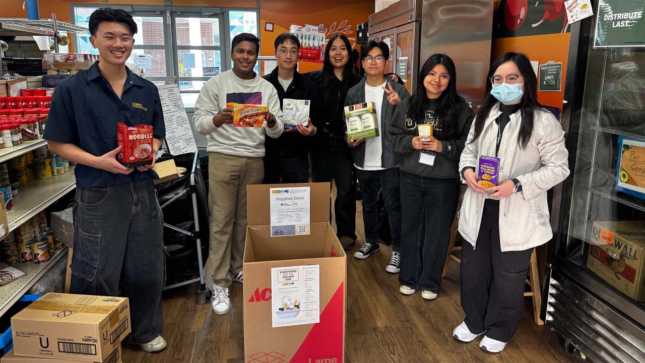 A group of seven people holding food and hygenic products near a donation box.