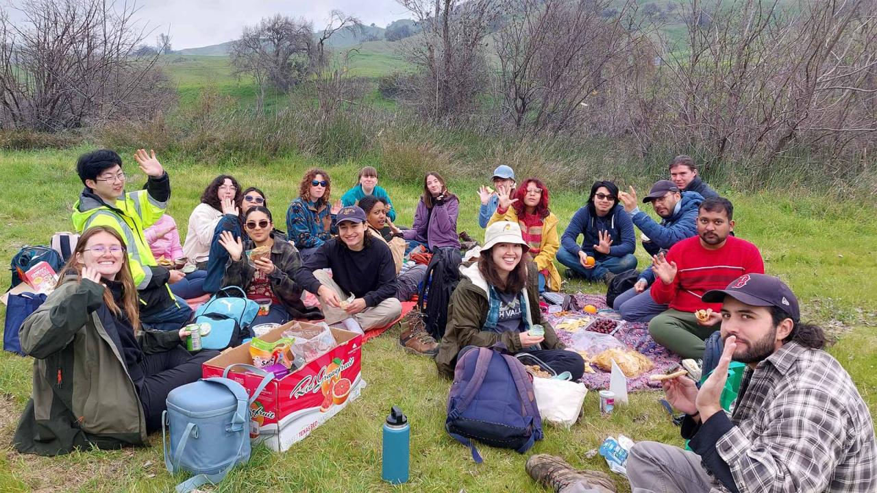 Diverse group of about 20 people picnicking on blankets on a green hillside under gray sky.