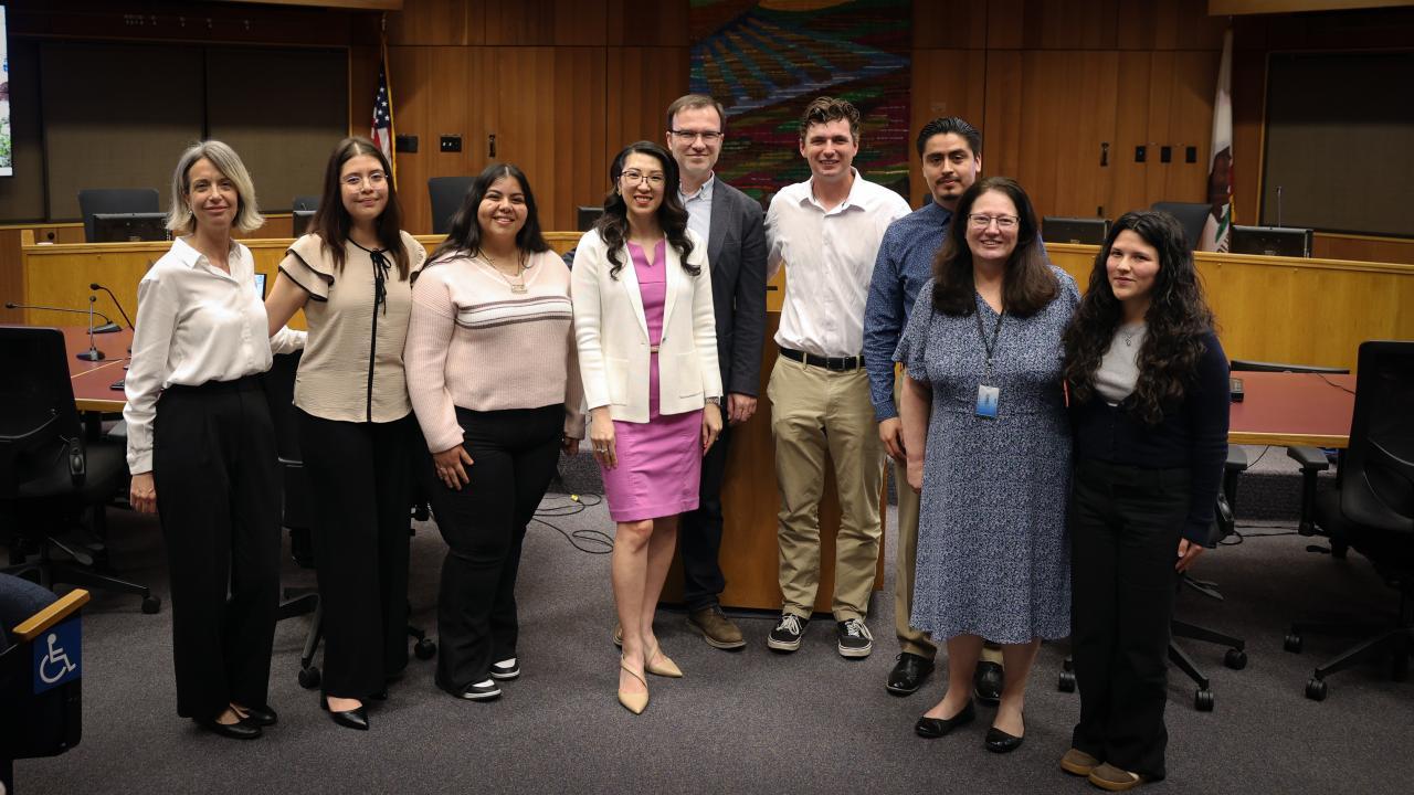Group photo of ten people in a wood-paneled council room, smiling in business attire
