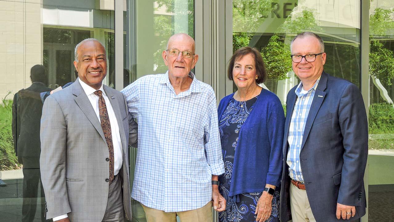 Four smiling adults standing together outside a modern glass building.