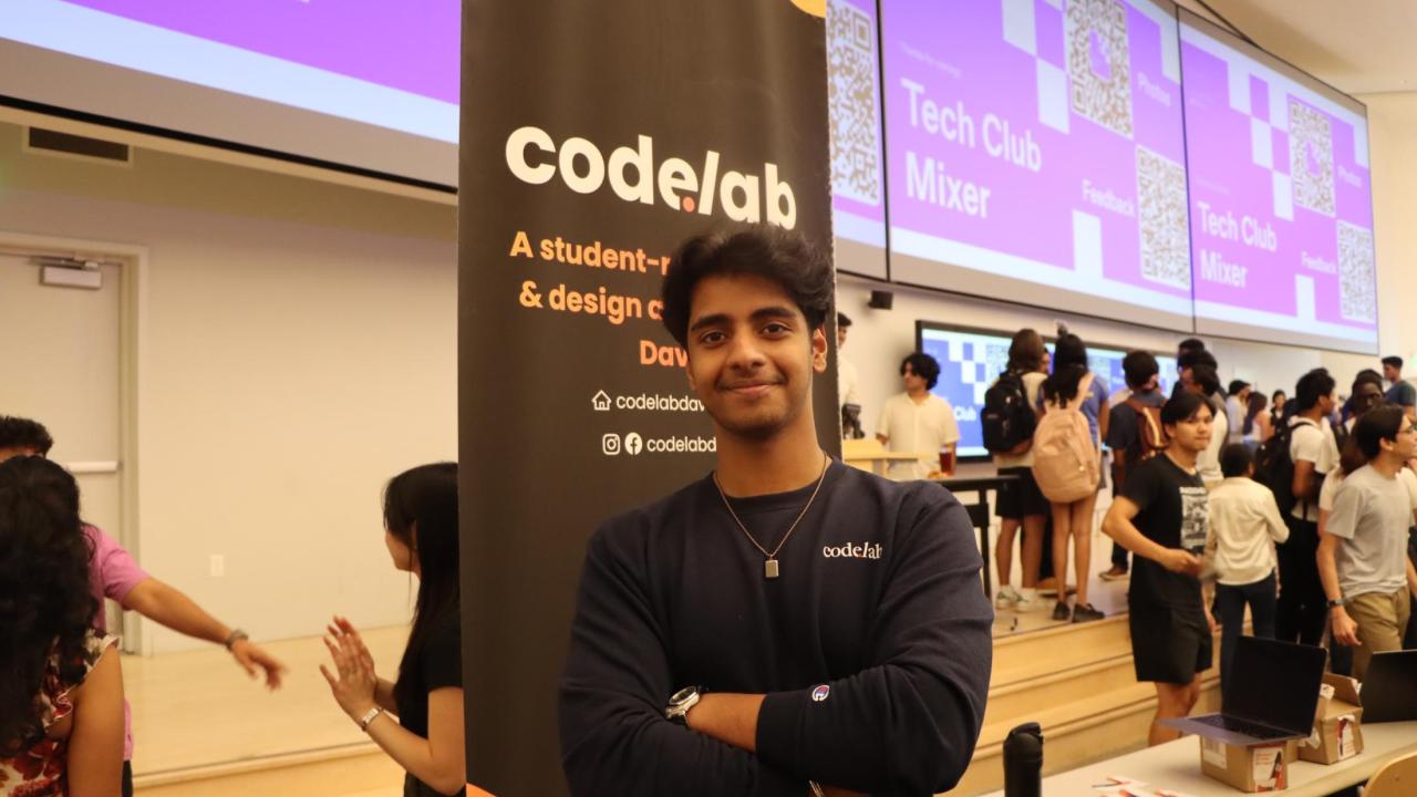 A student in a black long sleeve T-shirt, stands in front of a black banner that reads “code lab”, in a lecture hall filled with other students. 