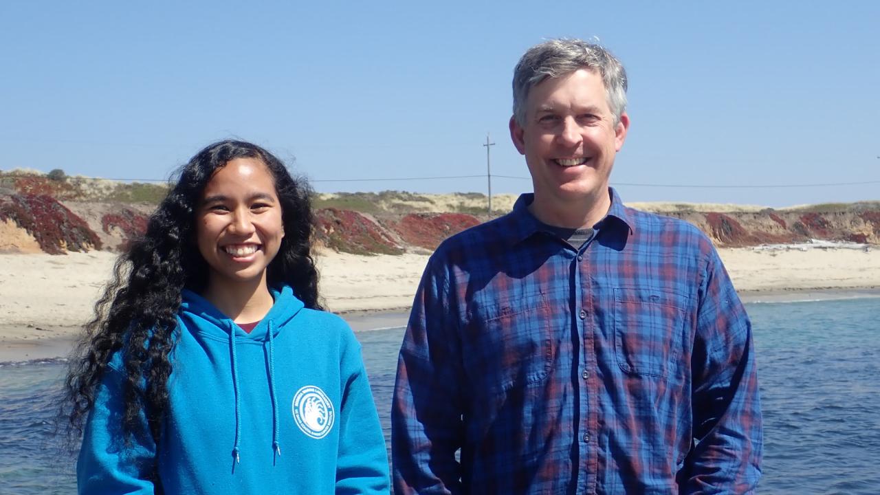 Jacquie Rajerison (left) and Professor Eric Sanford (right) at the Bodega Marine Reserve standing in front of the water and sandy coast. 