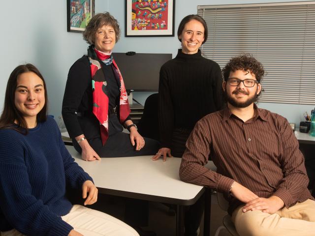 Four people smiling in an office setting, standing and sitting around a desk.