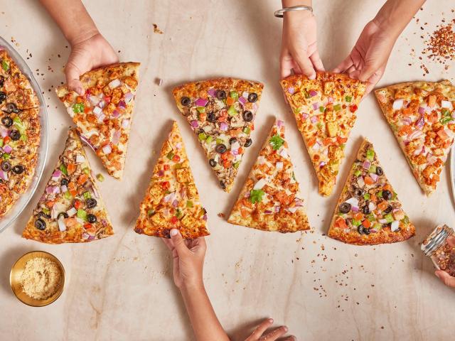 Hands reaching for slices of colorful pizza on a table with two pizza trays.
