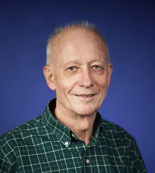 Smiling older man with gray hair, wearing a green checkered shirt, against a blue background.