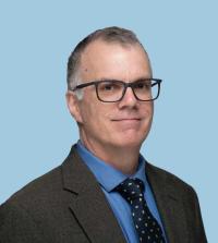 Headshot photograph of man with glasses, gray hair, suit, blue shirt, light blue background.