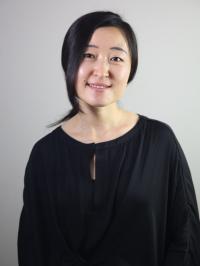Woman with dark hair dressed in dark clothing against gray background