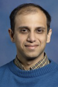 Headshot of a man in a blue sweater and checkered shirt, slight smile, blue background