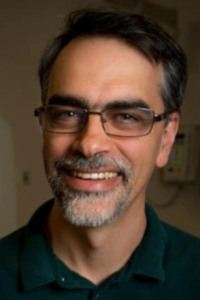 Smiling man with glasses, dark hair and grey beard wearing a black dress shirt