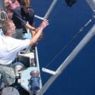 Photo of man on boat gesturing toward lake as people watch.