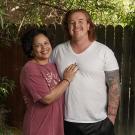 Portrait of a man and a woman standing together under trees and in front of a fence. He wears a white t-shirt and she wears and maroon t-shirt.