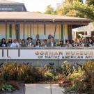 Group of people on a museum terrace behind a stone sign, mid-century building and gardens