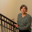 Woman in green patterned blouse on indoor staircase, holding railing and smiling towards the camera.