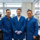 Three scientists in blue lab coats stand smiling in a laboratory filled with equipment.