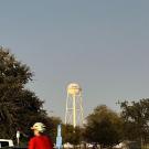 The UC Davis water tower rises above trees as a bicyclist rides past cars on a campus street at sunset.