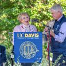 Four people at UC Davis podium outdoors; woman laughing as man speaks, university seal.