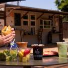 In an outdoor cafe setting, an unknown person is holding a sandwich above a table full of iced coffees, iced teas, salads and other food options. It's a sunny day with a blue sky peeking above the near food kiosk. 