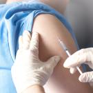 Gloved hands holding a needle and syringe against the bared upper arm of a person wearing a light blue top. 