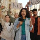people holding protest signs in background as woman with microphone stands in front