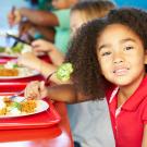 Child with dark curly hair and red shirt eats a school lunch. 